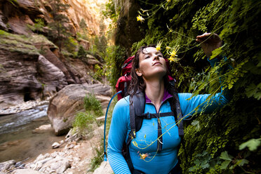 Eine Frau in einer blauen Basisschicht hält an, um während einer Rucksacktour durch die Narrows im Zion National Park in Springdale, Utah, an Blumen zu riechen. - AURF02075