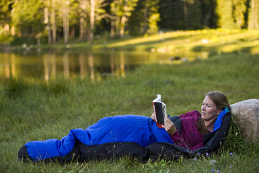A woman reads in a sleeping bag beside a mountain lake. - AURF02106