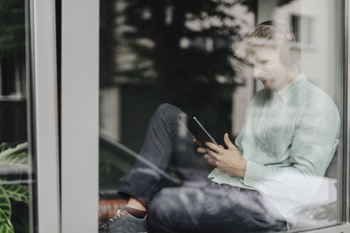 Junger Mann sitzt am Fenster und hört Musik mit Kopfhörern und digitalem Tablet - GUSF01228