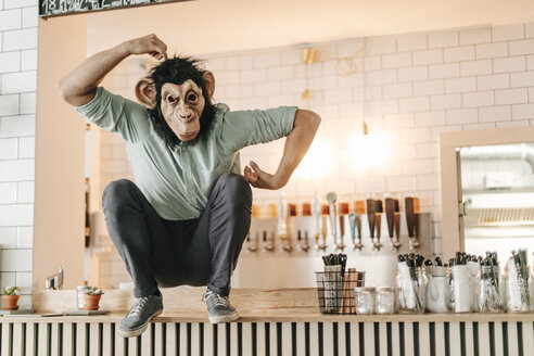 Man wearing monkey mask, sitting on counter of a bar, scratching head - GUSF01233