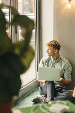 Junger Mann sitzt auf dem Boden, benutzt einen Laptop und schaut aus dem Fenster, lizenzfreies Stockfoto