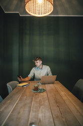 Mann sitzt im Büro, arbeitet bis spät in sein Start-up-Unternehmen, benutzt Laptop und Smartphone - GUSF01269