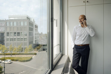 Smiling businessman standing in his office, talking on his mobile phone - KNSF04427