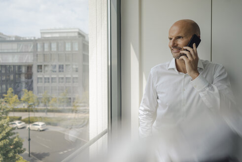 Smiling businessman standing in his office, talking on his mobile phone - KNSF04428