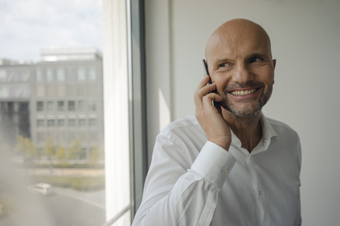Ein lächelnder Geschäftsmann steht in seinem Büro und telefoniert mit seinem Handy, lizenzfreies Stockfoto