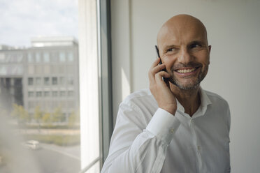 Ein lächelnder Geschäftsmann steht in seinem Büro und telefoniert mit seinem Handy - KNSF04429