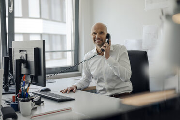 Smiling businessman sitting at his desk, talking on the phone - KNSF04435