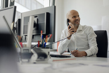 Smiling businessman sitting at his desk, talking on the phone - KNSF04437