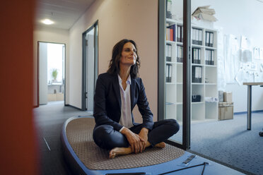 Businesswoman sitting cross-legged on paddle board, daydreaming in office - KNSF04499