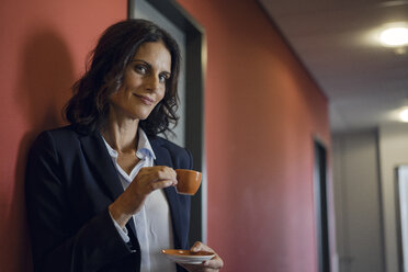 Mature businesswoman standing in office corridor, drinking coffee - KNSF04505