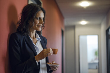 Mature businesswoman standing in office corridor, drinking coffee - KNSF04506