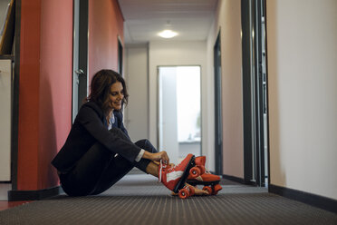 Mature businesswoman sitting in office corridor, putting on roller skates - KNSF04507