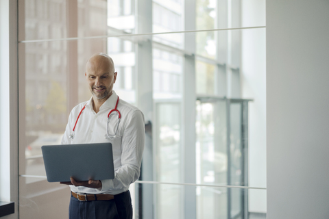 Doctor standing in hospital with stethoscope around his neck, holding laptop stock photo