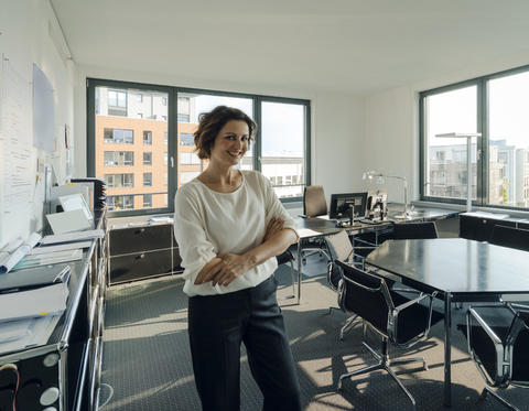 Erfolgreiche Geschäftsfrau steht mit verschränkten Armen in ihrem Büro, lizenzfreies Stockfoto