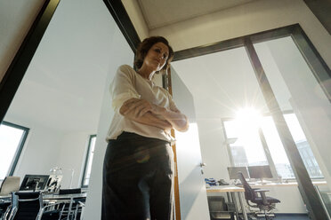 Businesswoman standing in office, with arms crossed, looking down - KNSF04552