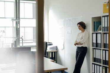 Businesswoman leaning against wall in office, with arms crossed - KNSF04555