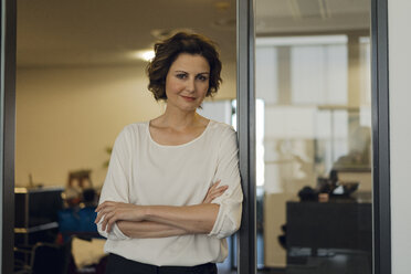 Successful businesswoman leaning in glass door, with arms crossed - KNSF04558