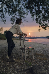 Woman leaning on a chair at the river, watching sunset - KNSF04581