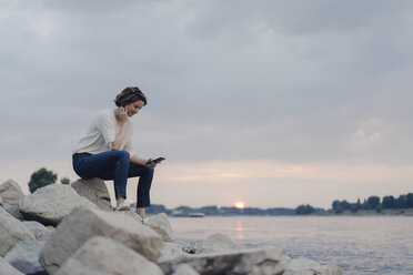 Woman sitting at the river, using smartphone - KNSF04586