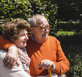 Senior couple sitting on bench in a park, with arms around - UUF14932