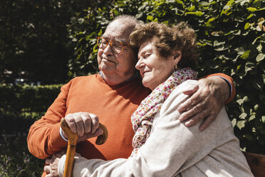 Senior couple sitting on bench in a park, with arms around - UUF14933