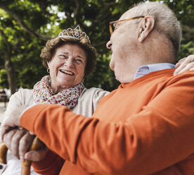 Happy senior couple sitting in park, woman wearing crown - UUF14954