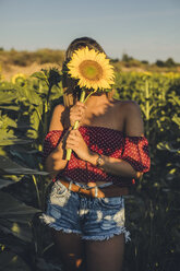 Junge Frau steht auf einem Feld und hält sich eine Sonnenblume vor das Gesicht - ACPF00296