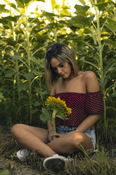 Junge Frau sitzt in einem Sonnenblumenfeld und hält eine Sonnenblume in der Hand - ACPF00299