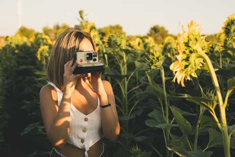 Junge Frau in einem Sonnenblumenfeld beim Fotografieren mit einer Sofortbildkamera, lizenzfreies Stockfoto