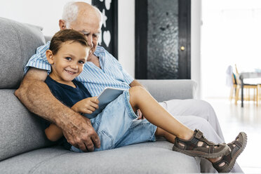 Porträt eines zufriedenen kleinen Jungen mit digitalem Tablet, der neben seinem Großvater auf der Couch zu Hause sitzt - JRFF01802