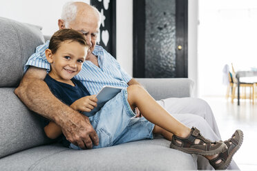 Portrait of content little boy with digital tablet sitting besides his grandfather on the couch at home - JRFF01802