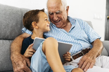 Happy little boy with digital tablet sitting beside his grandfather on the couch at home having fun - JRFF01803