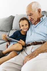 Portrait of happy little boy with digital tablet sitting besides his grandfather on the couch at home - JRFF01807
