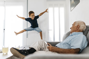 Großvater sitzt mit Tablet auf dem Sofa und beobachtet seinen Enkel, der in die Luft springt - JRFF01814