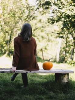 Rückenansicht einer Frau, die auf einer Holzbank neben einem Kürbis sitzt - RAMAF00061