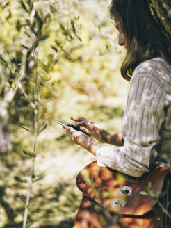 Italy, woman leaning against olive tree using cell phone - RAMAF00071