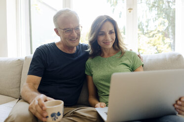 Lächelndes reifes Paar sitzt auf der Couch zu Hause und teilt sich einen Laptop - KNSF04597