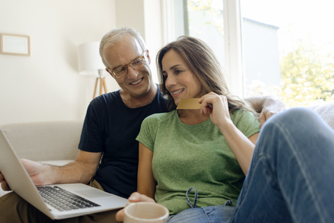 Lächelndes reifes Paar sitzt auf der Couch zu Hause beim Online-Shopping mit Laptop, lizenzfreies Stockfoto