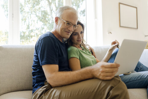 Smiling mature couple sitting on couch at home sharing tablet - KNSF04599