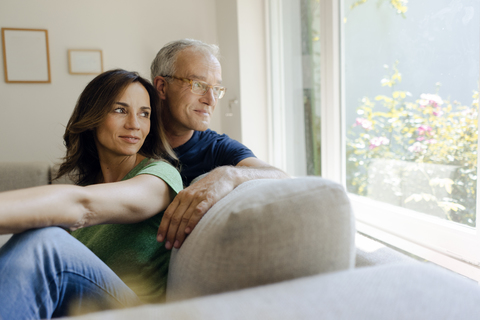 Lächelndes reifes Paar sitzt zu Hause auf der Couch und schaut aus dem Fenster, lizenzfreies Stockfoto
