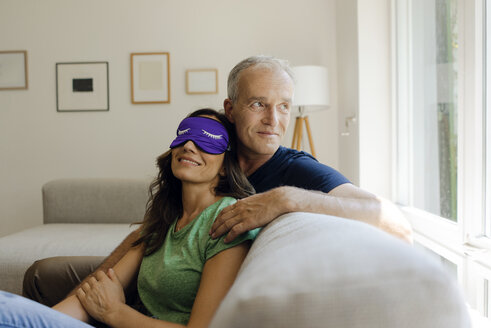 Smiling mature couple sitting on couch at home with woman wearing eye mask - KNSF04602