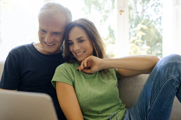 Smiling mature couple sitting on couch at home sharing laptop - KNSF04608