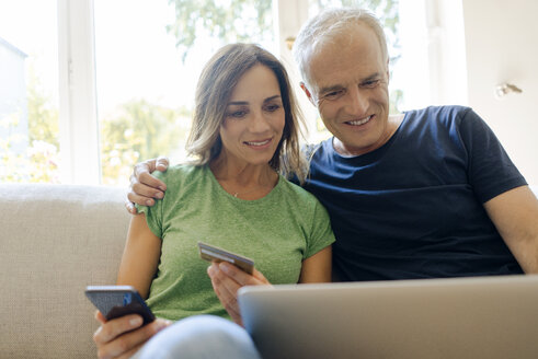 Smiling mature couple sitting on couch at home shopping online with laptop and smartphone - KNSF04609