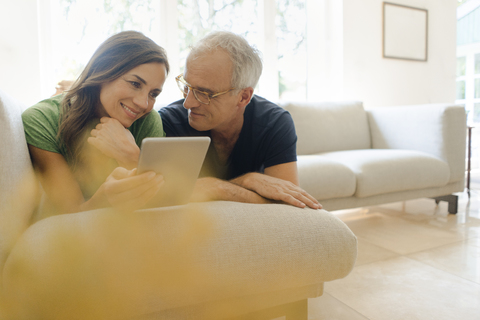 Lächelndes reifes Paar, das zu Hause auf der Couch liegt und ein Tablet teilt, lizenzfreies Stockfoto