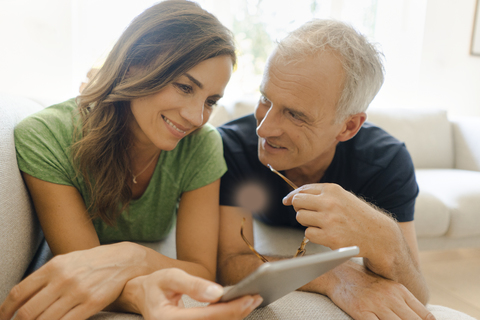 Lächelndes reifes Paar, das zu Hause auf der Couch liegt und ein Tablet teilt, lizenzfreies Stockfoto