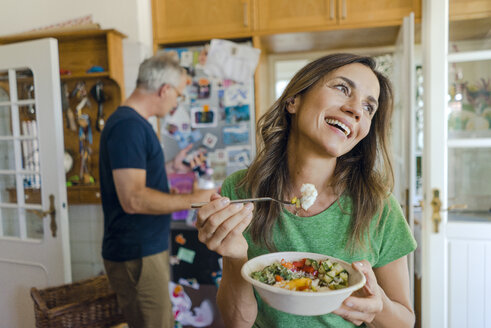 Glückliche reife Frau zu Hause isst einen Salat mit Mann im Hintergrund - KNSF04622