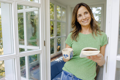 Lächelnde reife Frau zu Hause mit Buch und Kaffeetasse - KNSF04623
