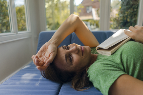 Smiling mature woman lying on couch at home with book stock photo