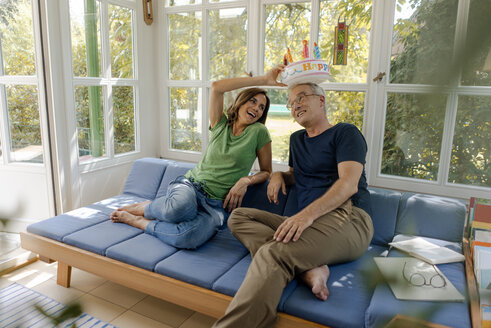 Verspielt reifes Paar sitzt auf Couch zu Hause mit gefälschten Geburtstagskuchen - KNSF04636