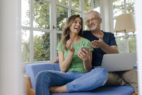 Happy mature couple sitting on couch at home with cell phone and laptop - KNSF04643